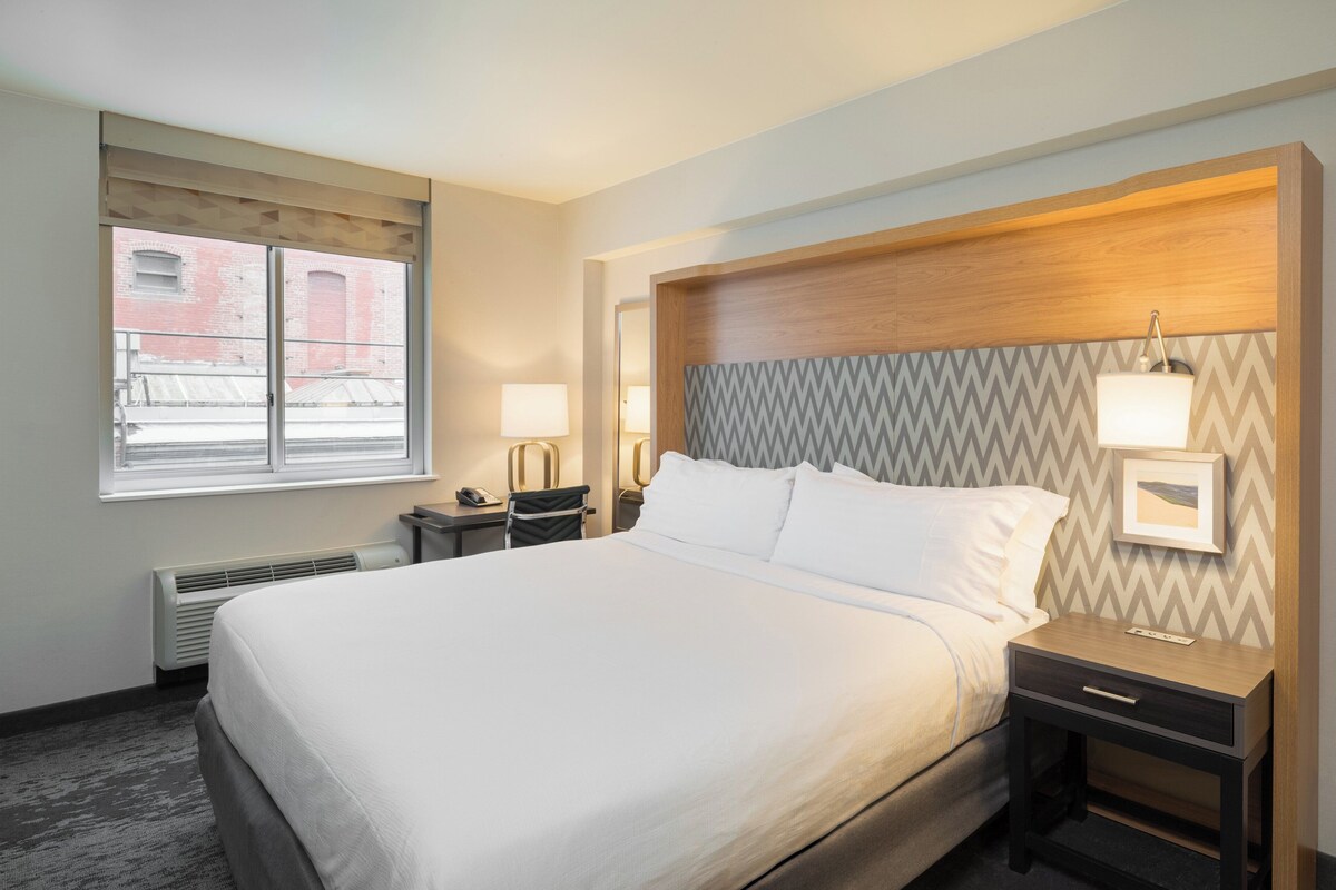 A hotel room with a large bed, two bedside tables, a window with a view of a brick building, and a wall with a zigzag pattern.