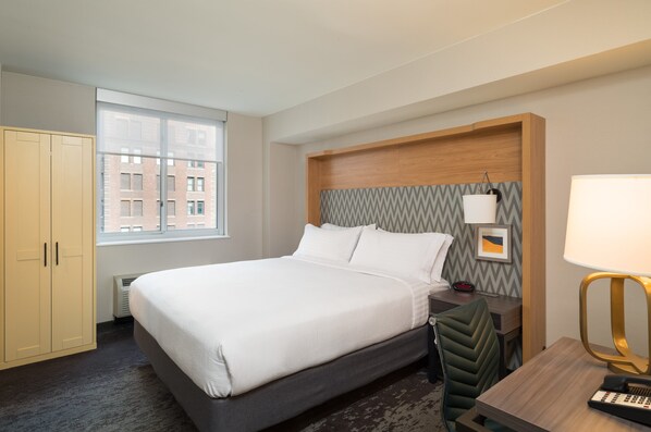 A hotel room with a bed, a desk, a chair, a lamp, a heater, and a wardrobe. - Holiday Inn New York City - Wall Street by IHG (New York)