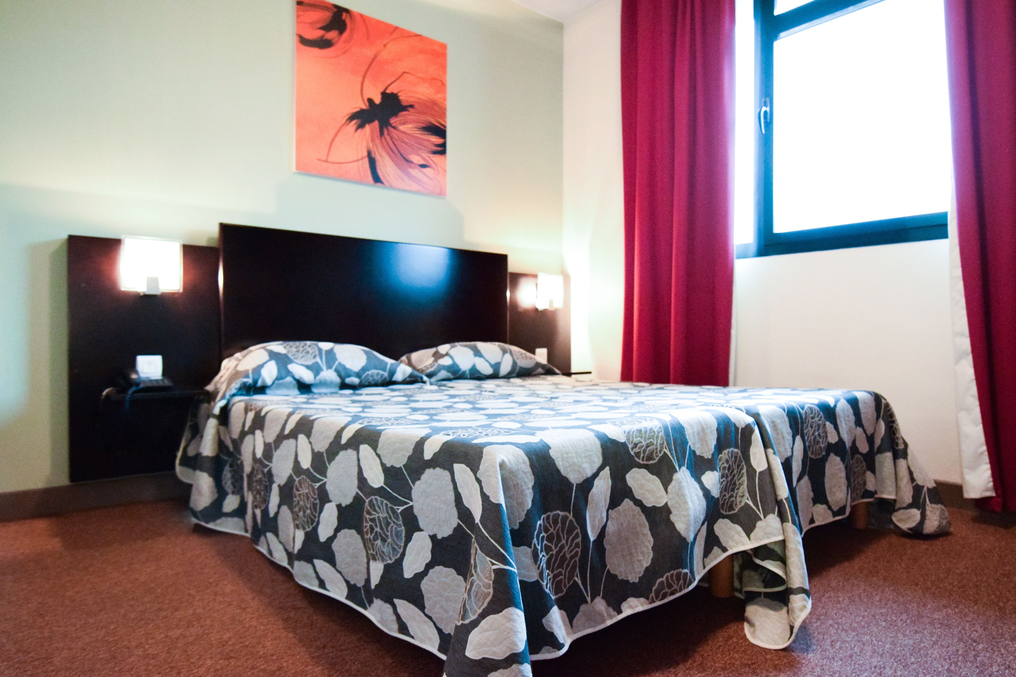 Une chambre d’hôtel avec un lit, une table de chevet, une lampe fixée au mur et une fenêtre avec des rideaux rouges.