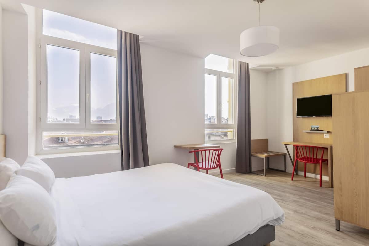 Ein Hotelzimmer mit einem großen Bett, einem Schreibtisch, einem Sessel, einem Fernseher und Blick auf die Stadt durch die Fenster.