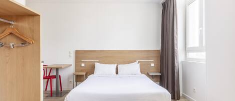 A hotel room with a bed, a wooden wardrobe, a desk with a chair, and a window with curtains.