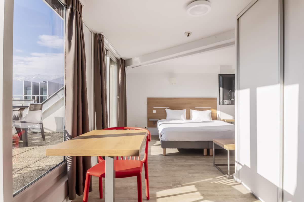 Ein Hotelzimmer mit Bett, Schreibtisch und Stuhl. Ein Fenster mit Vorhängen bietet Blick auf die Berge.