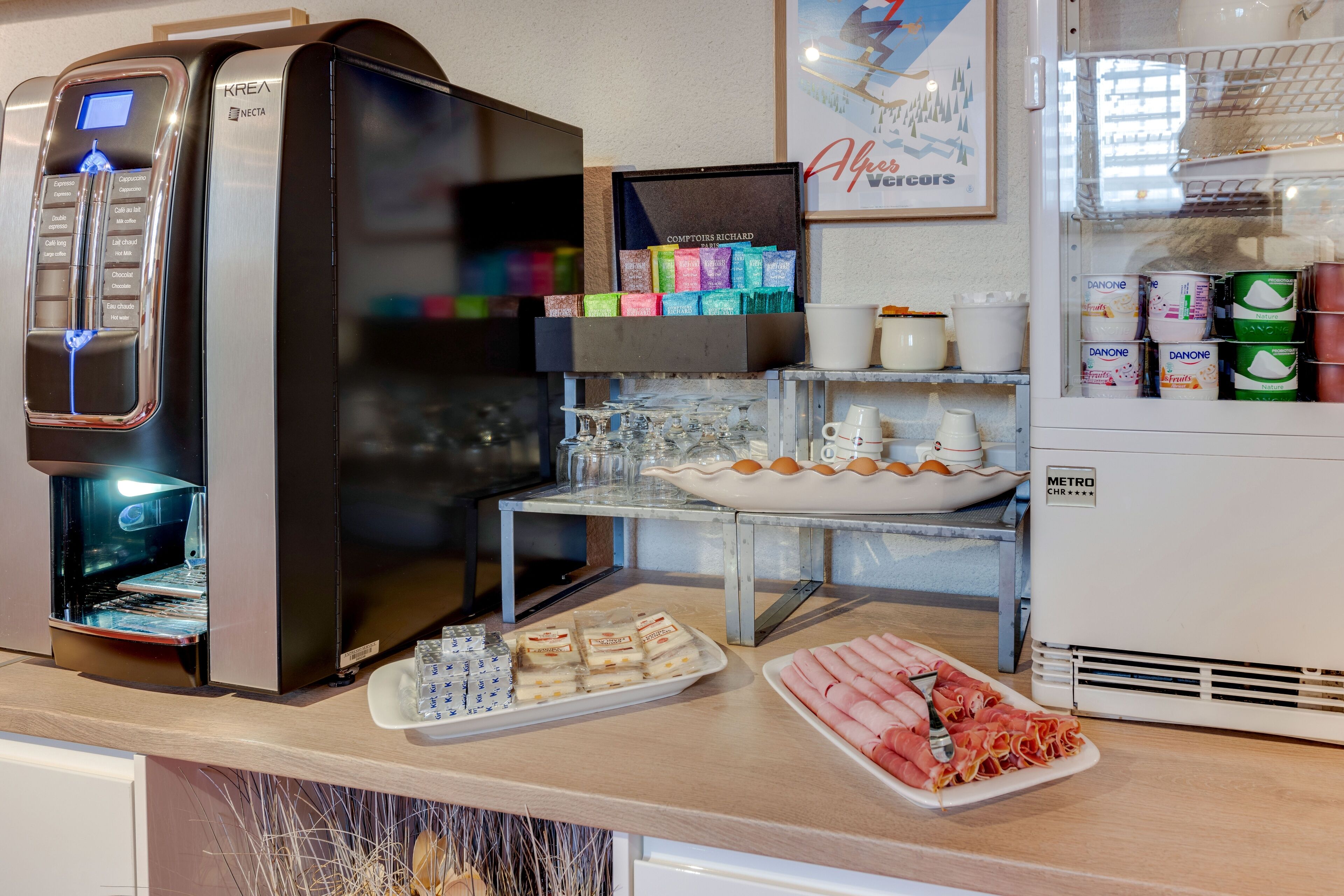 A breakfast bar with a coffee machine, a display of Danone yogurt cups, and a variety of pastries and meats.