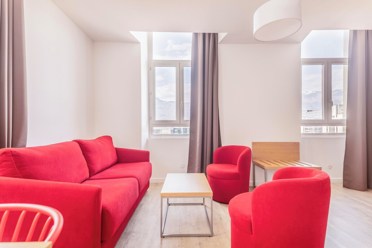 A modern living room with a red sofa, a wooden coffee table, and a red armchair. There are two windows with curtains, a white ceiling lamp, and a view of the cityscape.