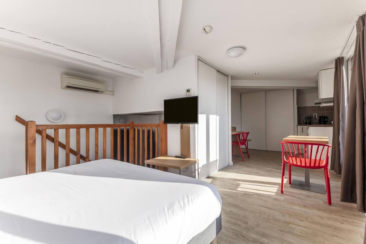 Ein modernes Hotelzimmer mit Bett, Fernseher, kleinem Tisch, rotem Sessel und einer kitchenette im Hintergrund.