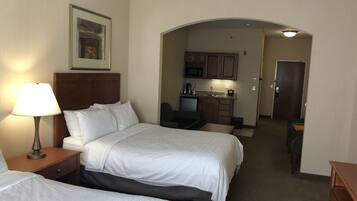 A hotel room with two beds, a desk, a chair, a lamp, a framed picture on the wall, and a kitchenette in the background.