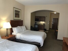 A hotel room with two beds, a desk, a chair, a lamp, a framed picture on the wall, and a kitchenette in the background.