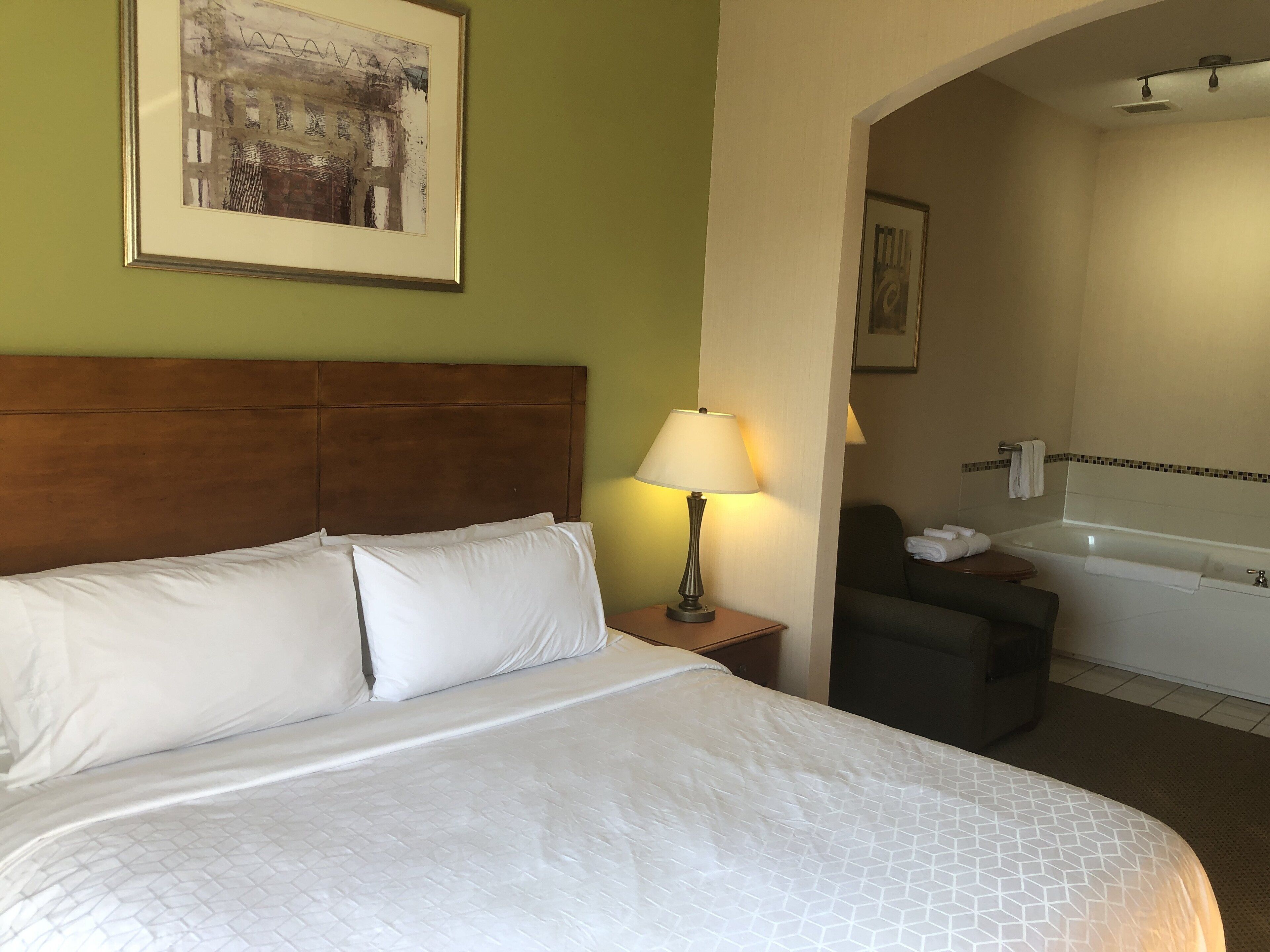 A hotel room with a large bed, a nightstand with a lamp, a chair, and a bathroom with a bathtub and towel rack.