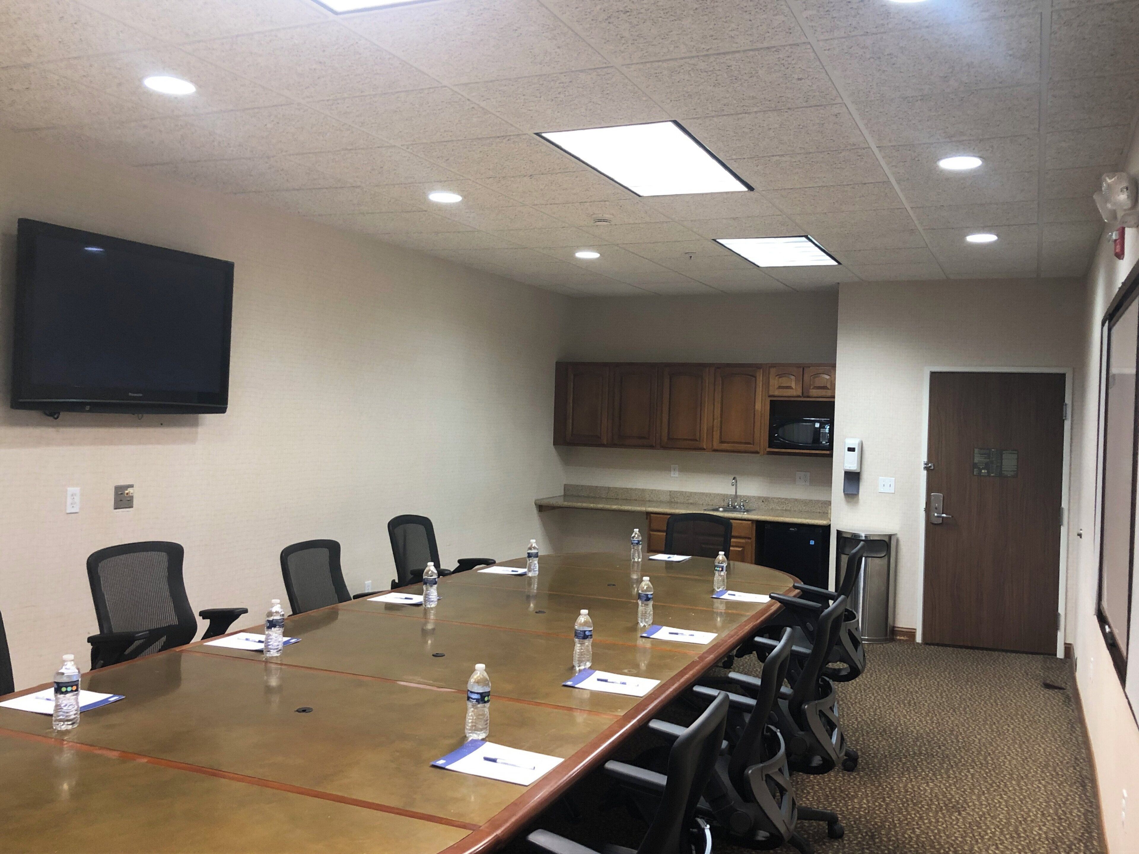 A conference room with a long table, chairs, water bottles, papers, a flat-screen TV, and a kitchenette in the background.