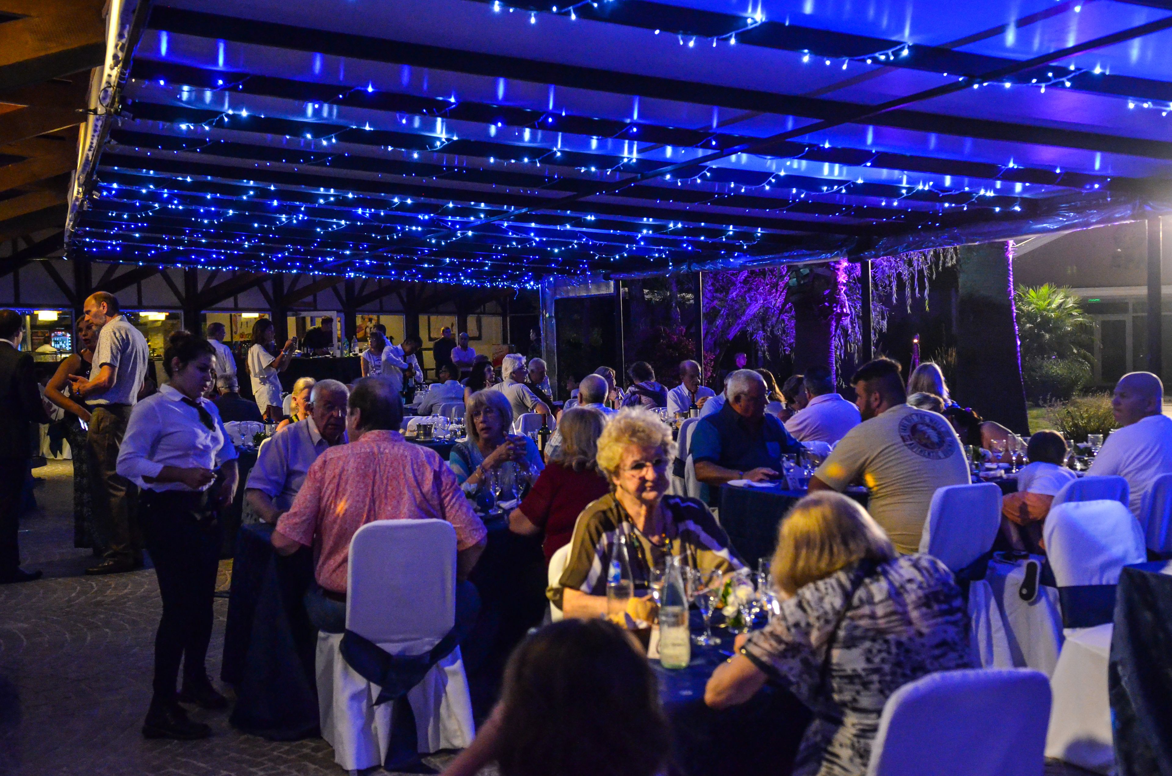 An outdoor dining area with tables and chairs, string lights overhead, and a crowd of people.