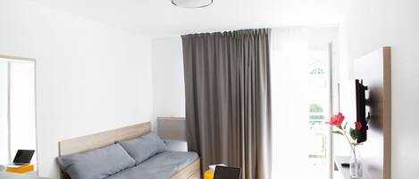 A modern bedroom with a bed, a laptop on a small table, and a window with curtains.