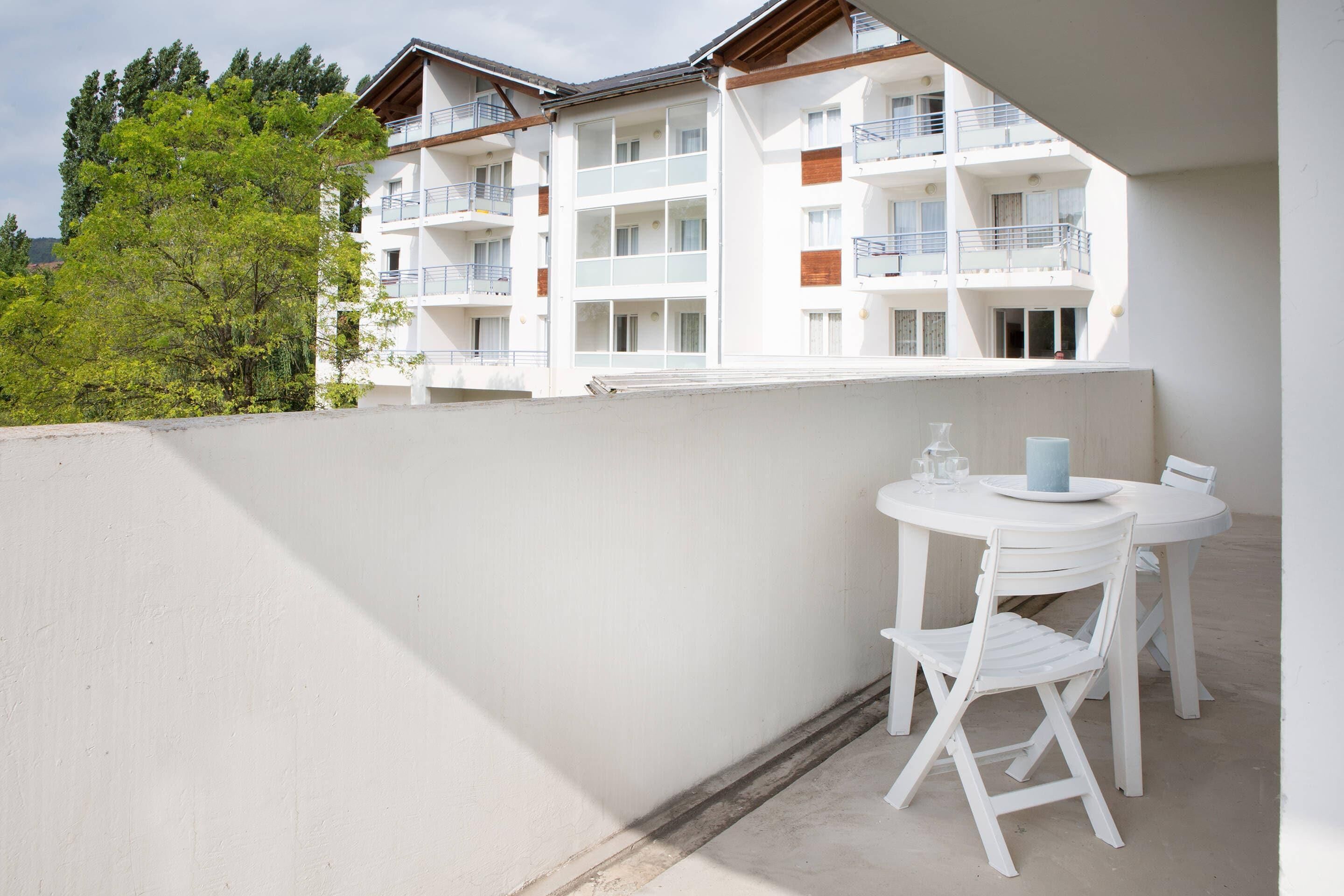 Un balcon blanc avec une table et des chaises, donnant sur un immeuble à plusieurs étages également doté de balcons.