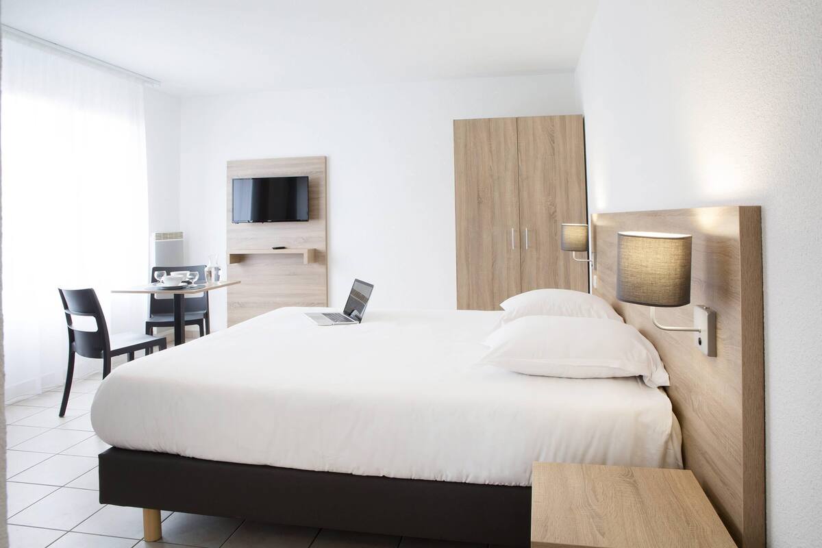 A modern hotel room with a large bed, a desk with a laptop, a TV mounted on the wall, and a bedside lamp.