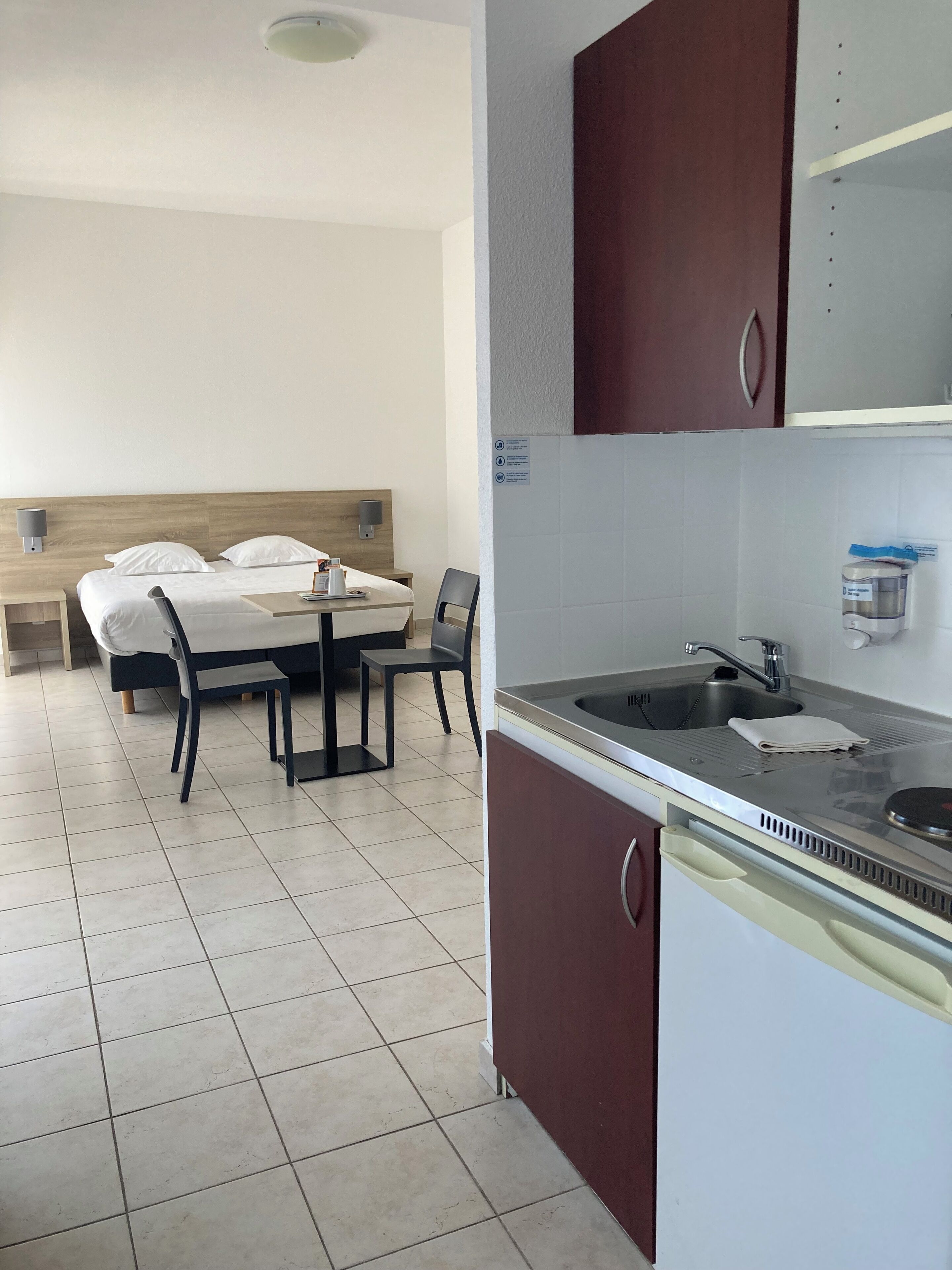 A hotel room with a bed, a dining table with chairs, a kitchen area with a sink and cabinets, and a ceiling light.