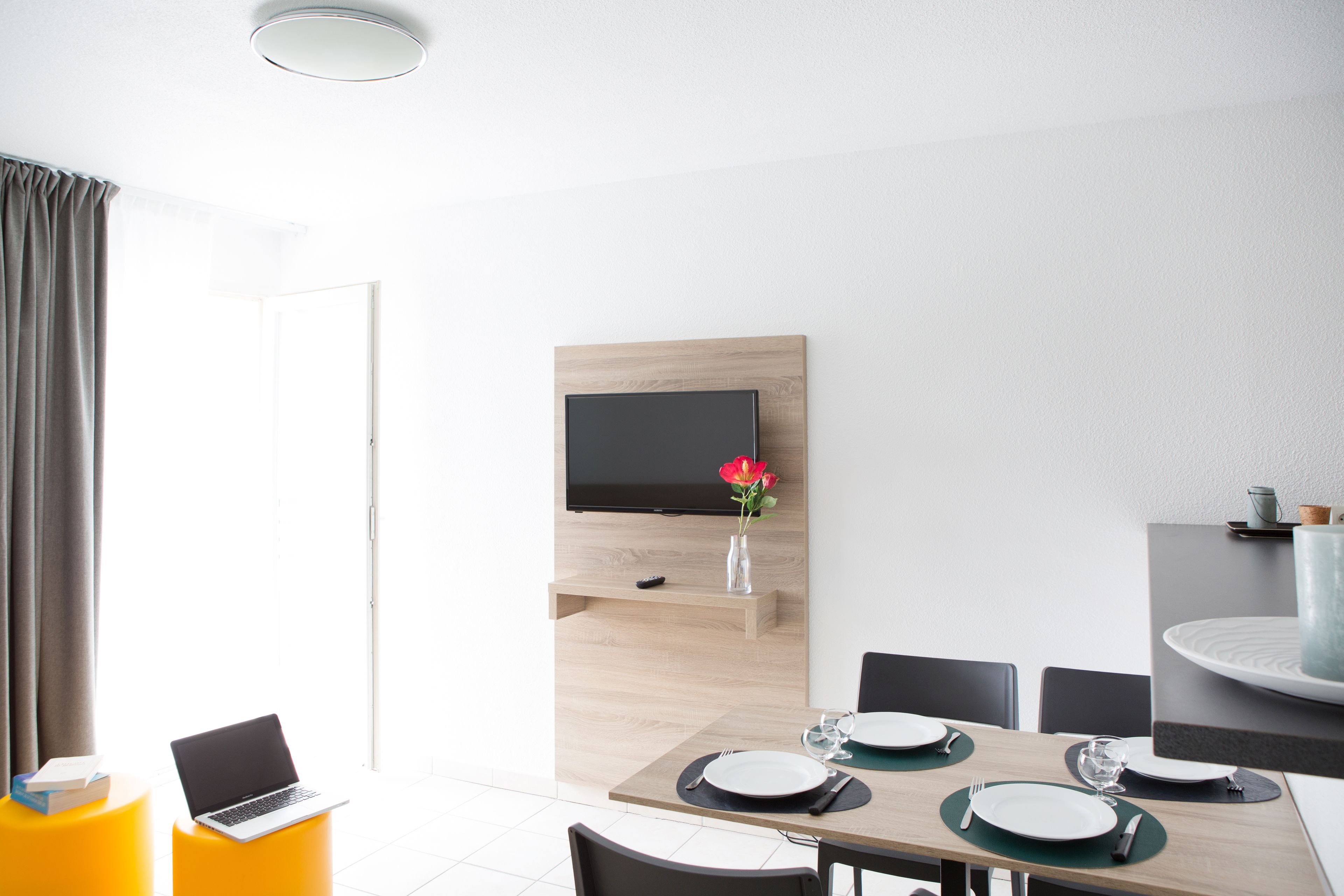 A modern dining area with a wooden table set for four, a flat-screen TV mounted on the wall, and a laptop on a yellow stool.