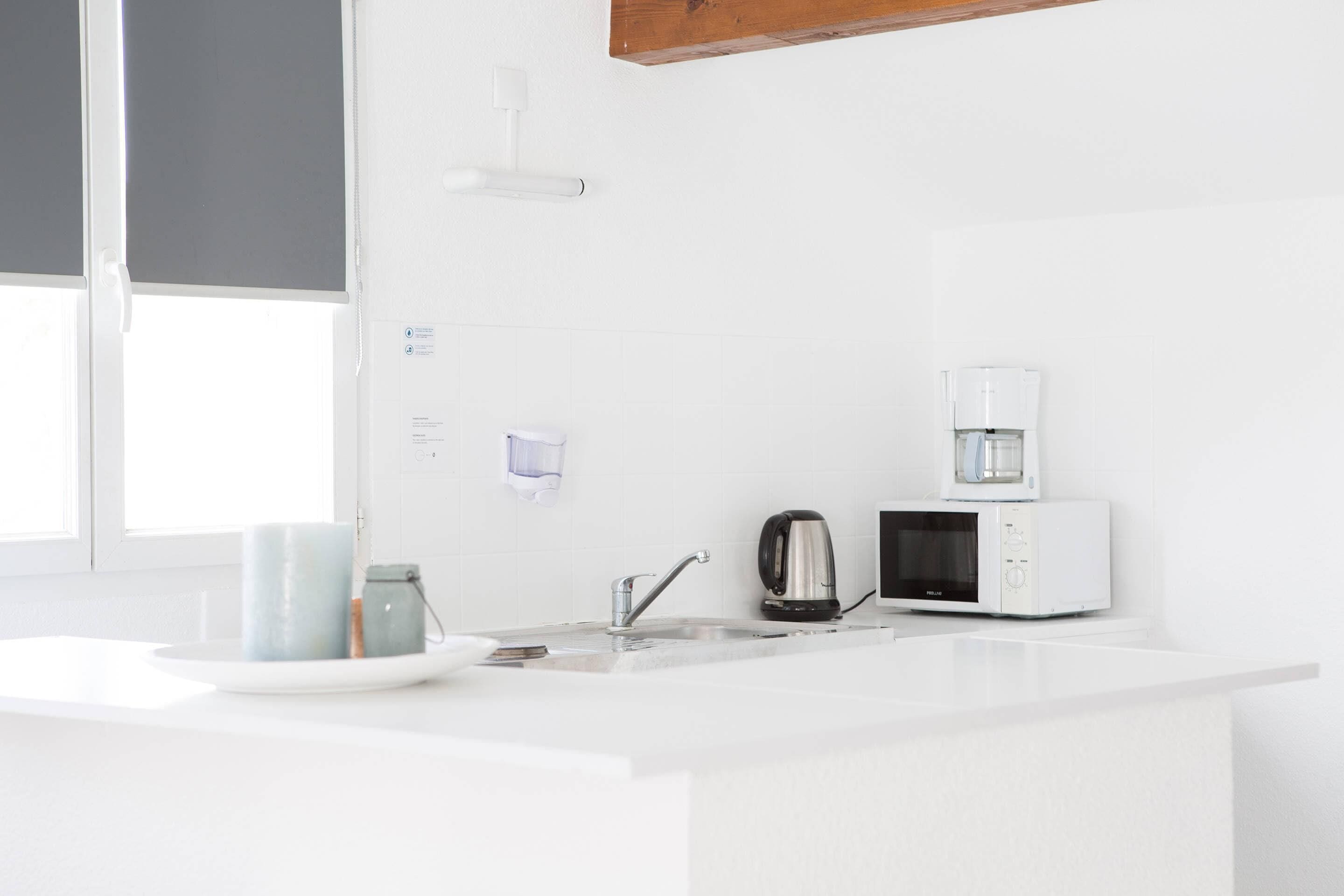 Une cuisine moderne dotée de plans de travail blancs, d’un évier en acier inoxydable, d’une bouilloire, d’une machine à café et d’un four à micro-ondes.