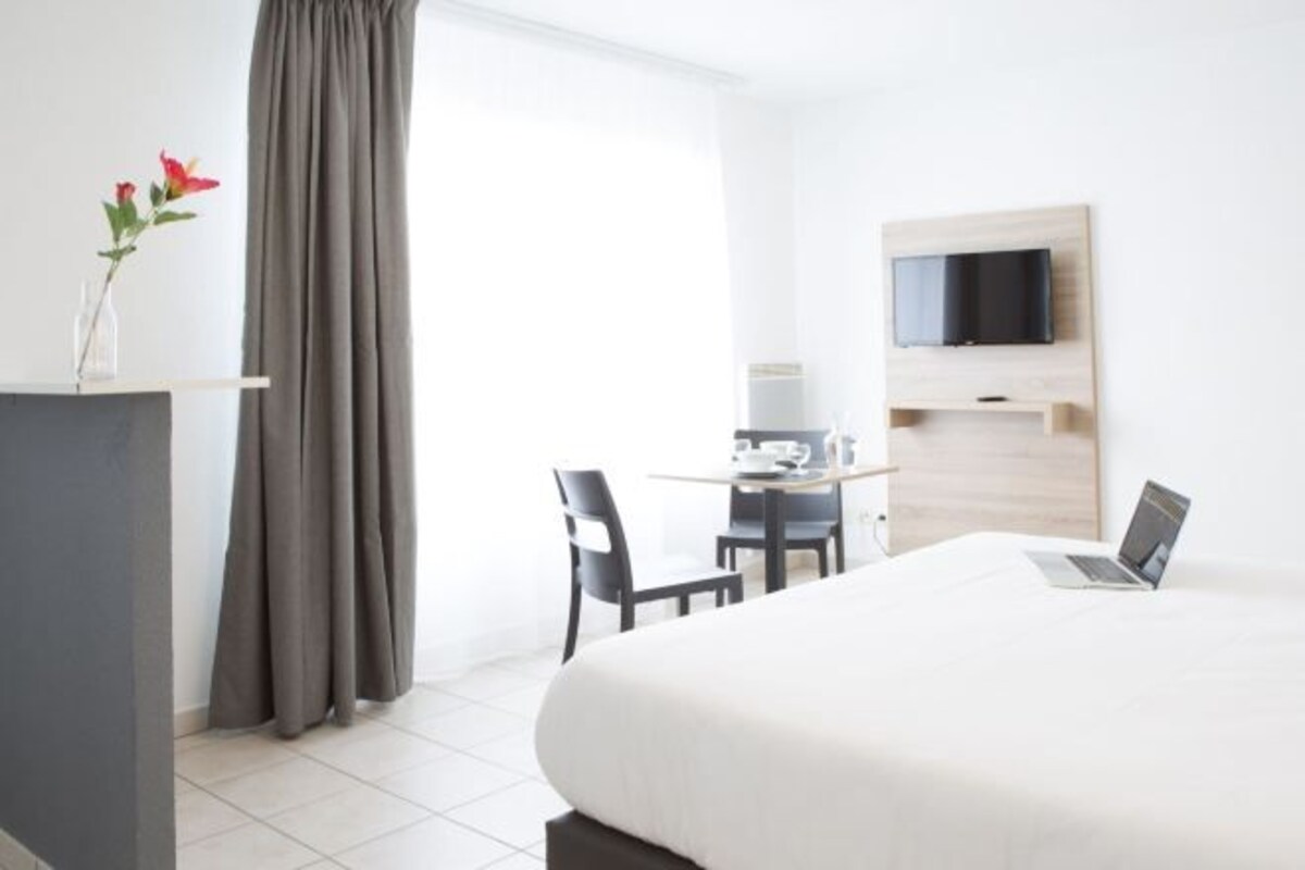 A hotel room with a bed, a desk with a laptop, a TV mounted on the wall, and a window with curtains.
