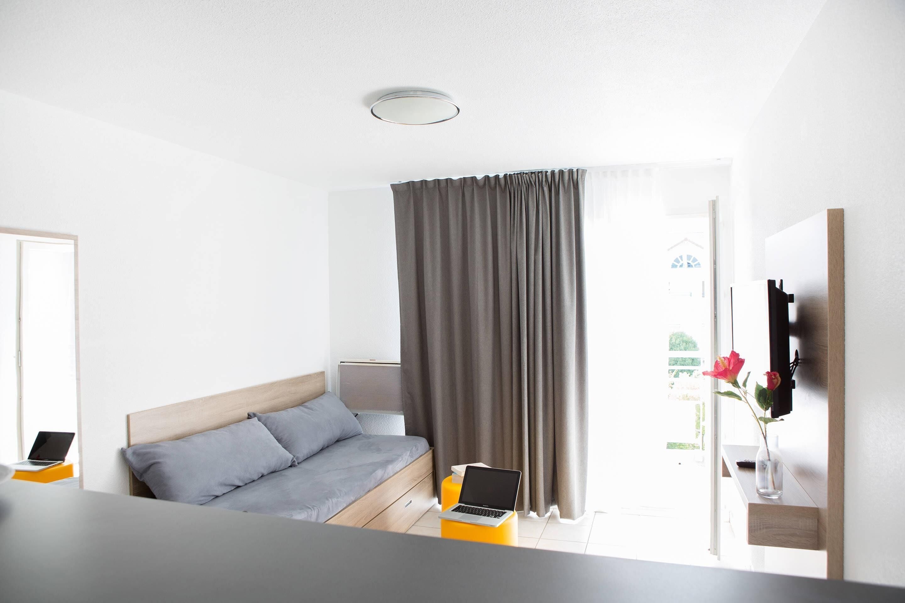 Ein modernes Hotelzimmer mit einem Bett, einem Laptop auf einem Beistelltisch, einer Vase mit Blumen und Blick ins Freie.