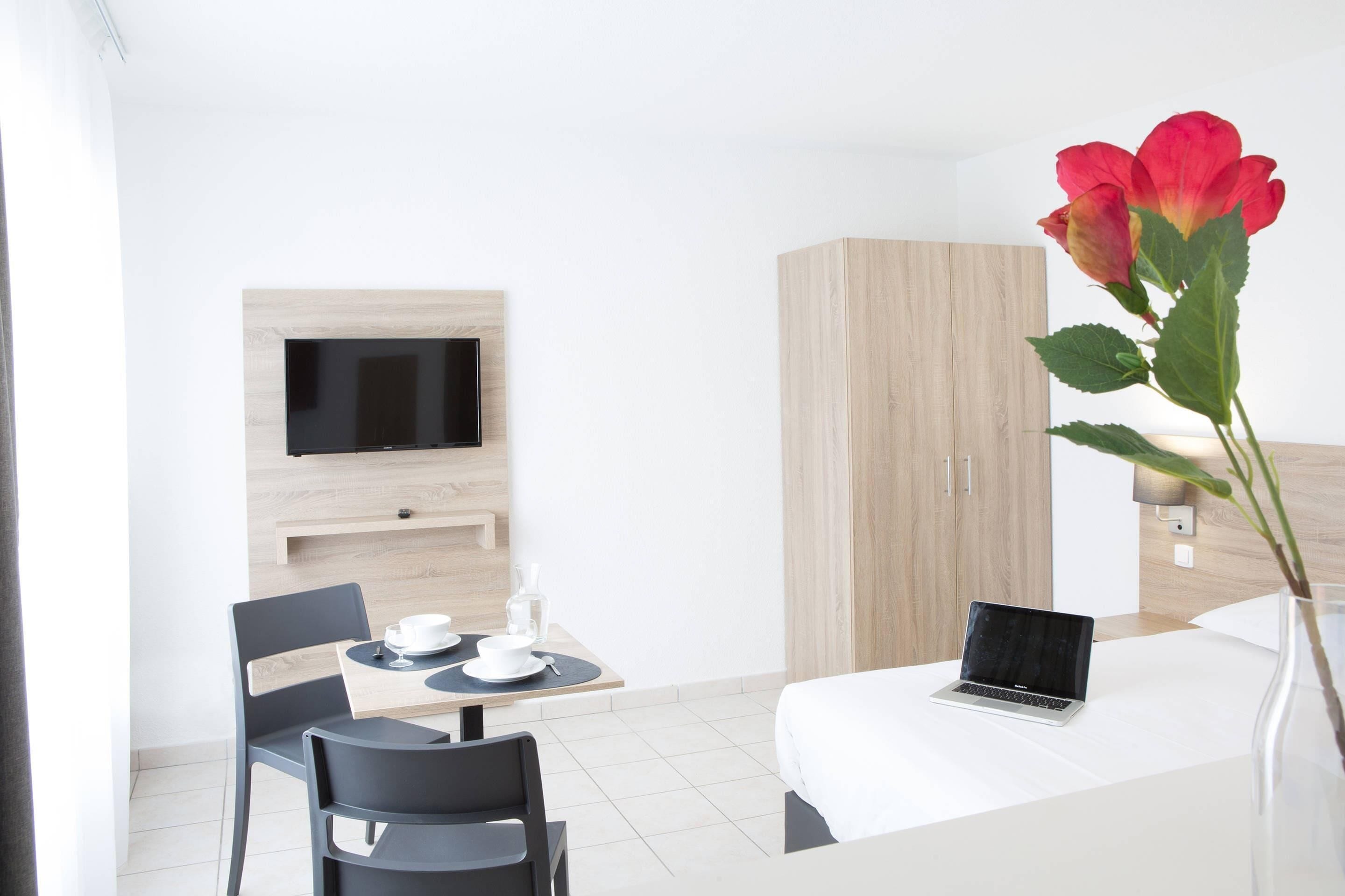 Une chambre d’hôtel moderne équipée d’un lit, d’un bureau avec un ordinateur portable, d’un téléviseur fixé au mur et d’un vase contenant une fleur rouge.