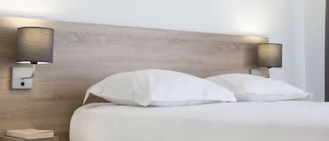 Una cama bien hecha con sábanas blancas y dos lámparas fijadas en la pared a cada lado.