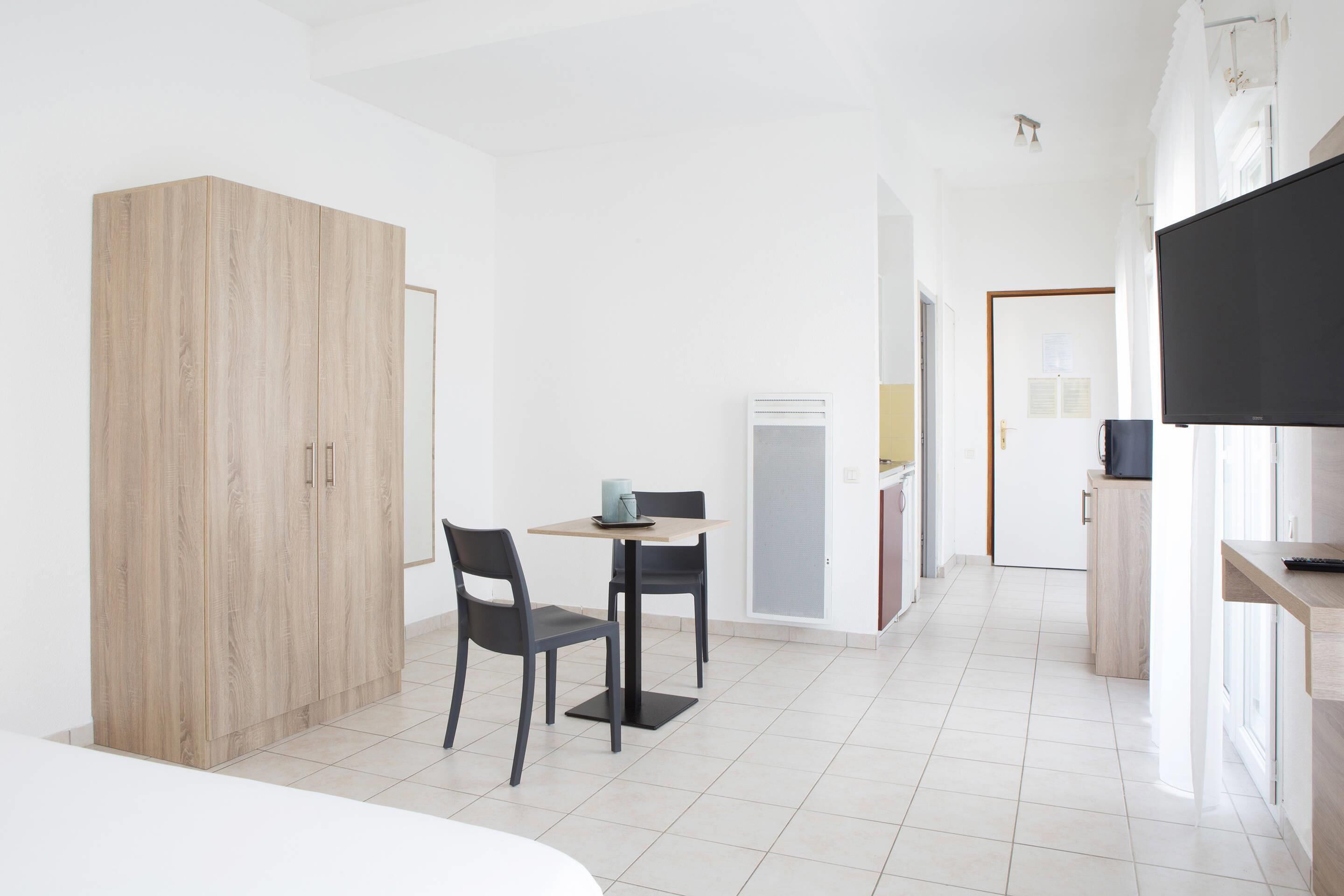 Une chambre moderne et minimaliste, avec un lit, une armoire en bois, une petite table à manger avec des chaises, un téléviseur et, au fond, un coin cuisine.