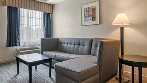 A modern hotel room with a grey sectional sofa, a small table, a floor lamp, and a framed picture on the wall. - Comfort Inn & Suites (Port Charlotte)