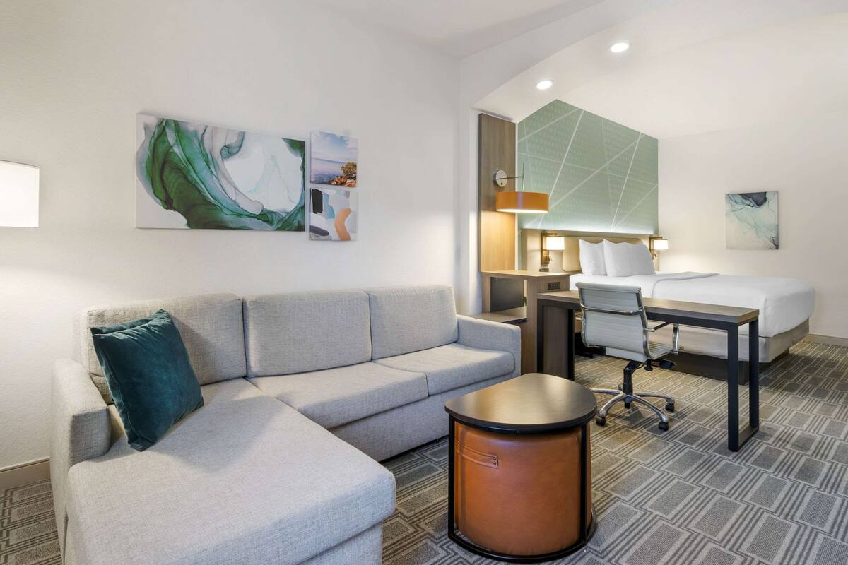 A modern hotel room with a grey sofa, a round coffee table, a desk with a chair, a bed, and abstract wall art.