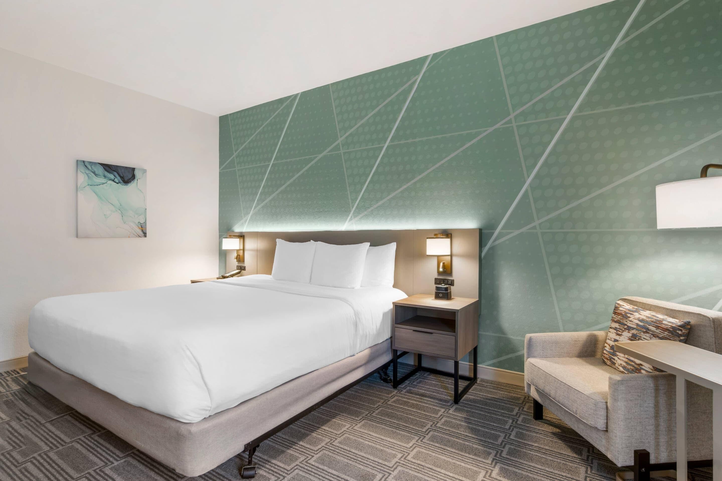 A hotel room with a bed, bedside table, lamp, and a small armchair.