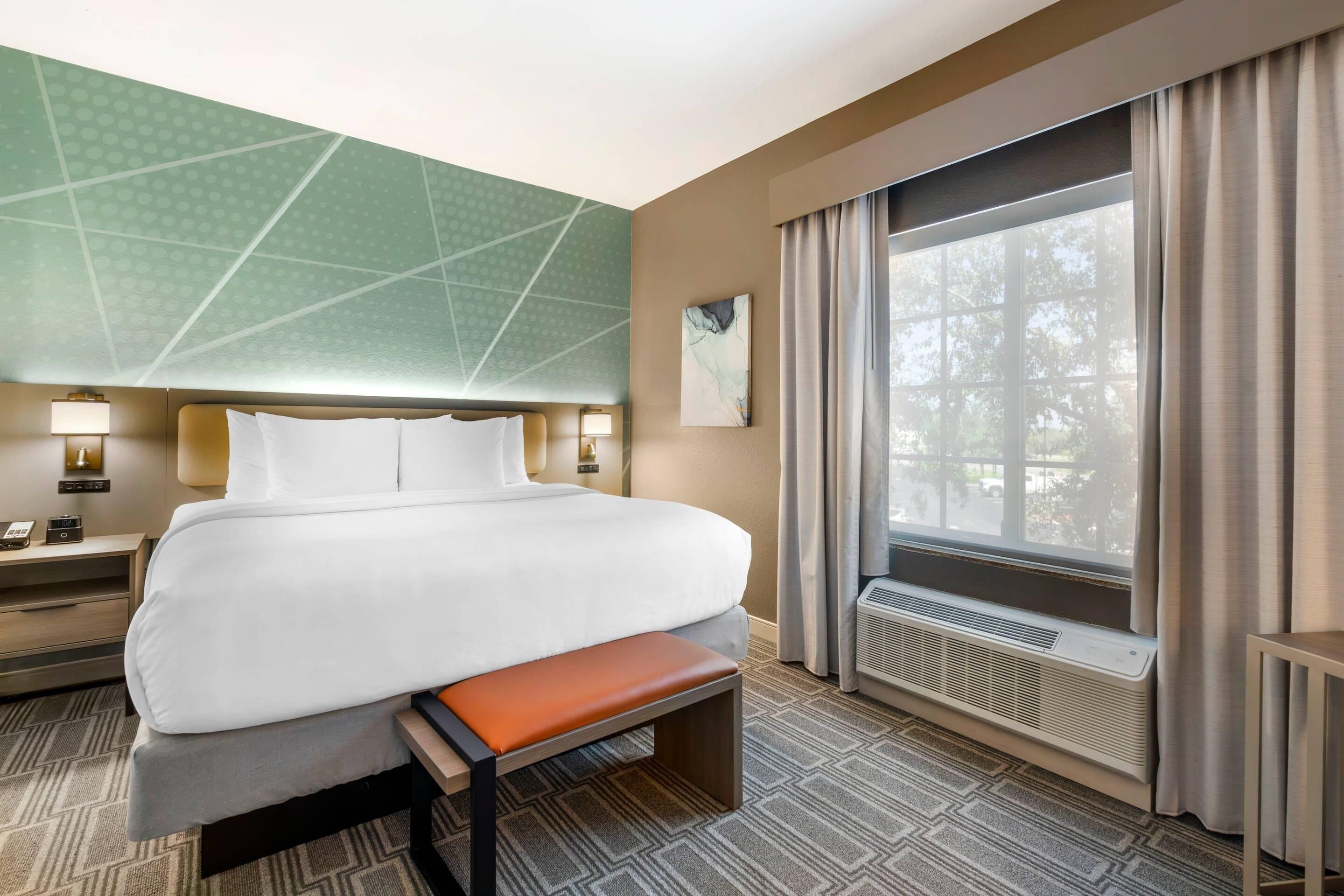 A hotel room with a bed, bedside tables, a bench, a large window with curtains, and a wall-mounted artwork.
