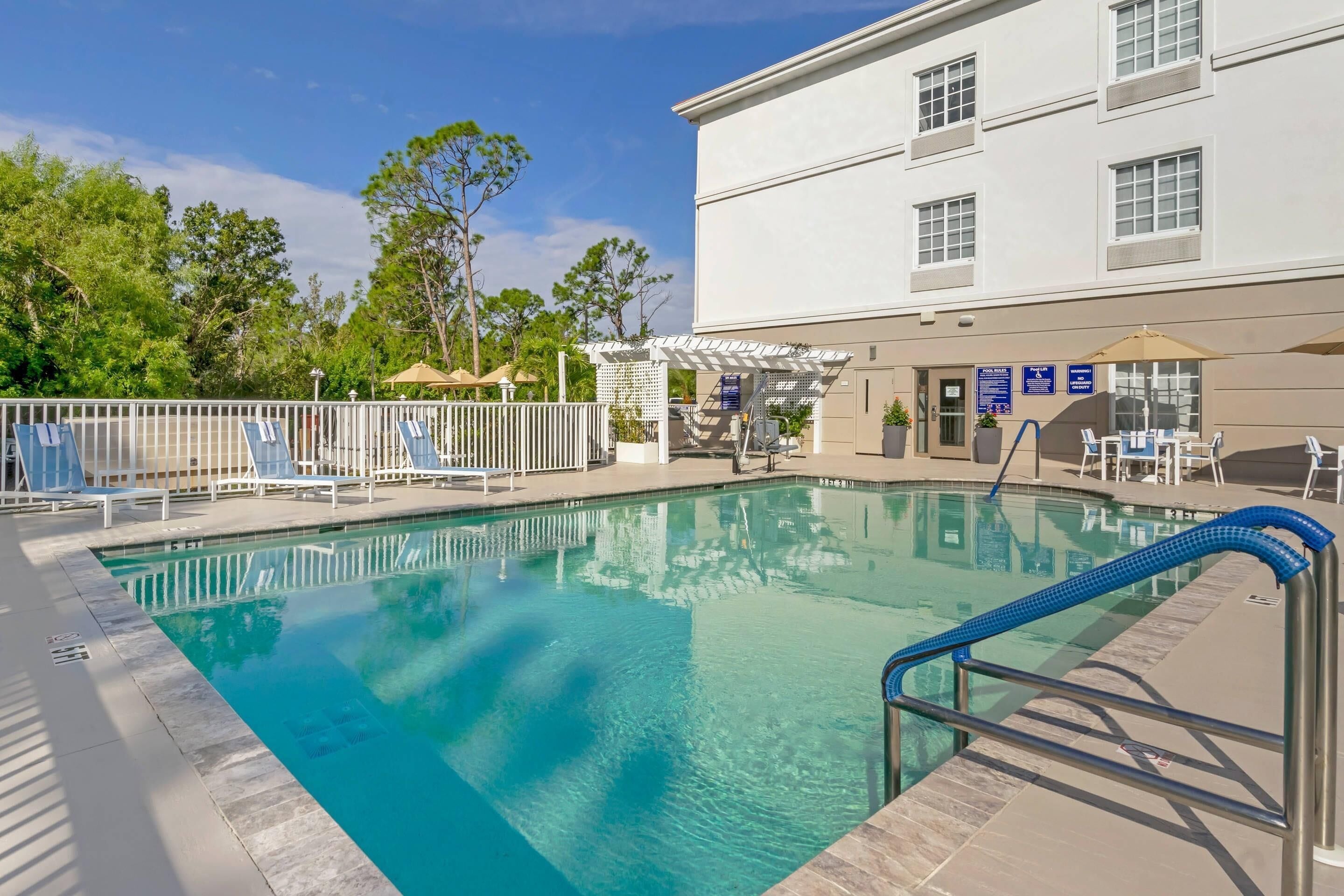 A clear swimming pool with a blue deck, surrounded by a white fence, and a building with multiple windows in the background.