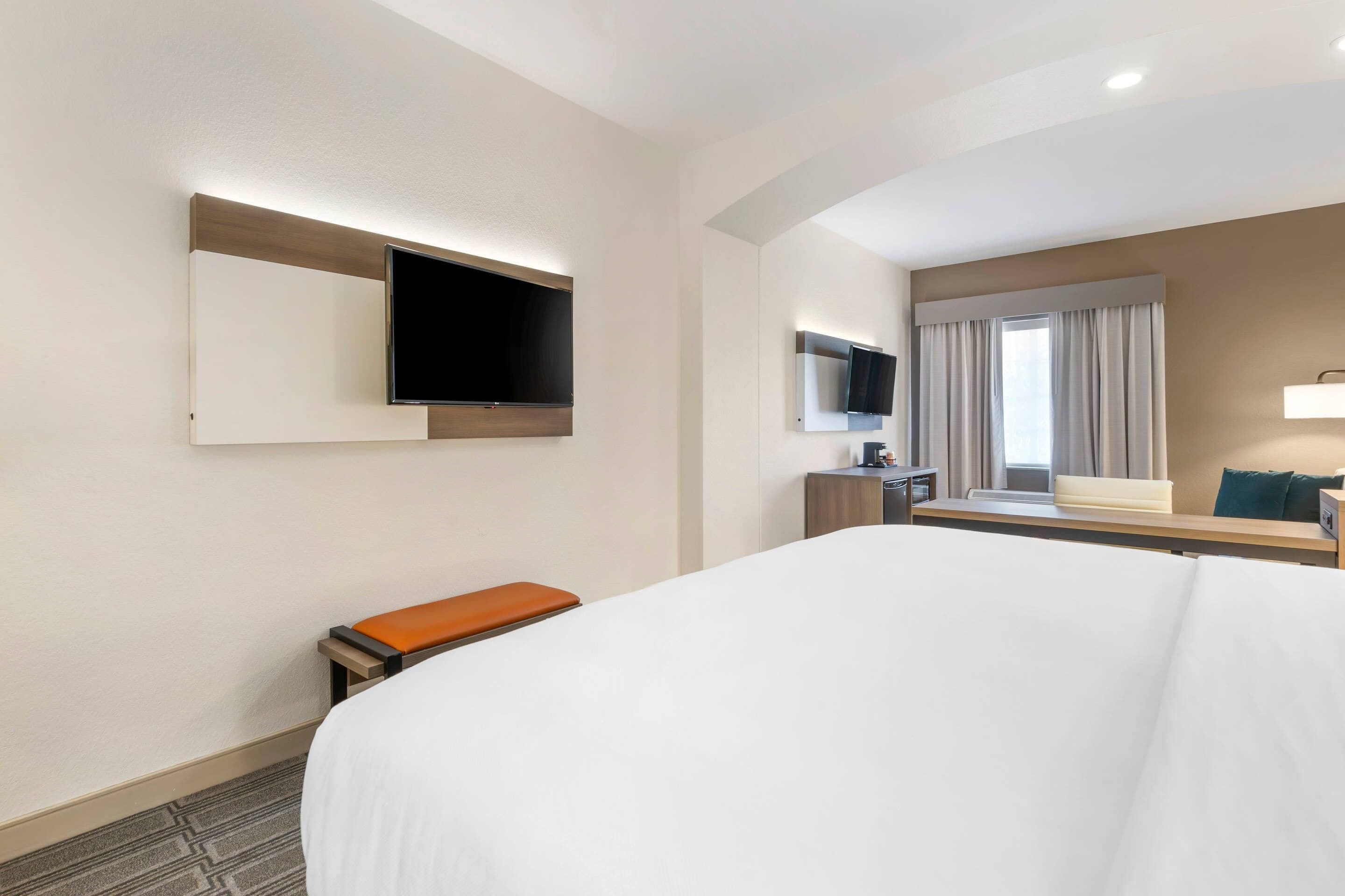 Une chambre d’hôtel avec un grand lit, un téléviseur à écran plat fixé au mur, un petit bureau avec une chaise et une fenêtre avec des rideaux.