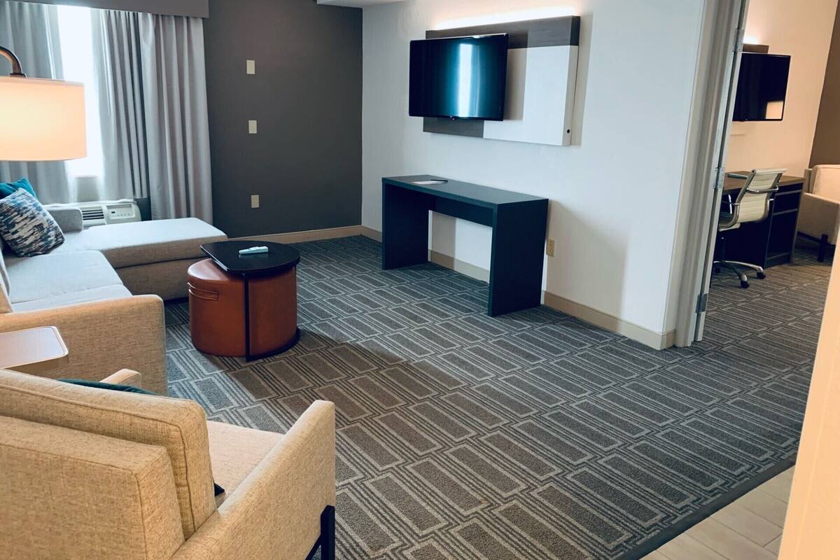 A hotel room with a sofa, a small round table, a desk, a flat-screen TV mounted on the wall, and a patterned carpet.