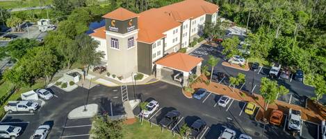 An aerial view of a hotel with a central tower, multiple parking lots, and surrounding greenery.