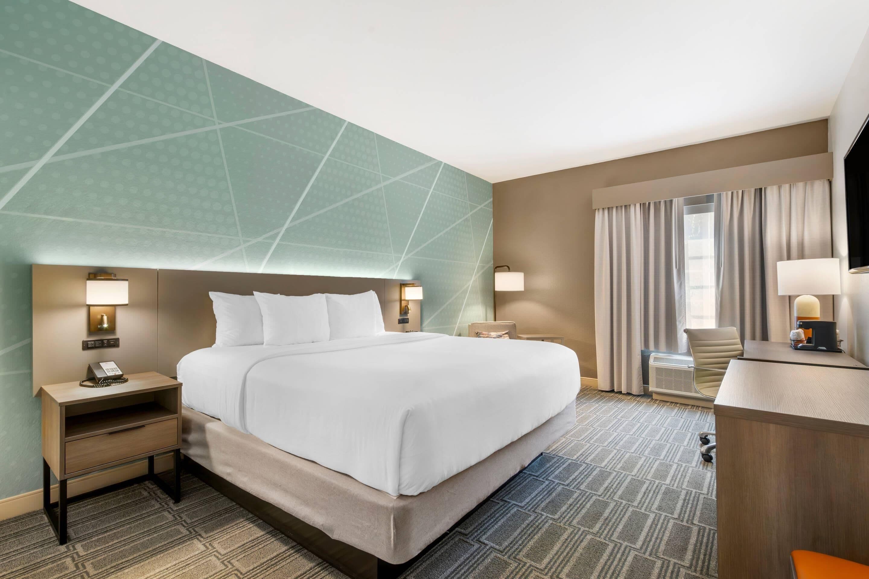 A hotel room with a large bed, bedside tables with lamps, a desk with a chair, and a large window with curtains.