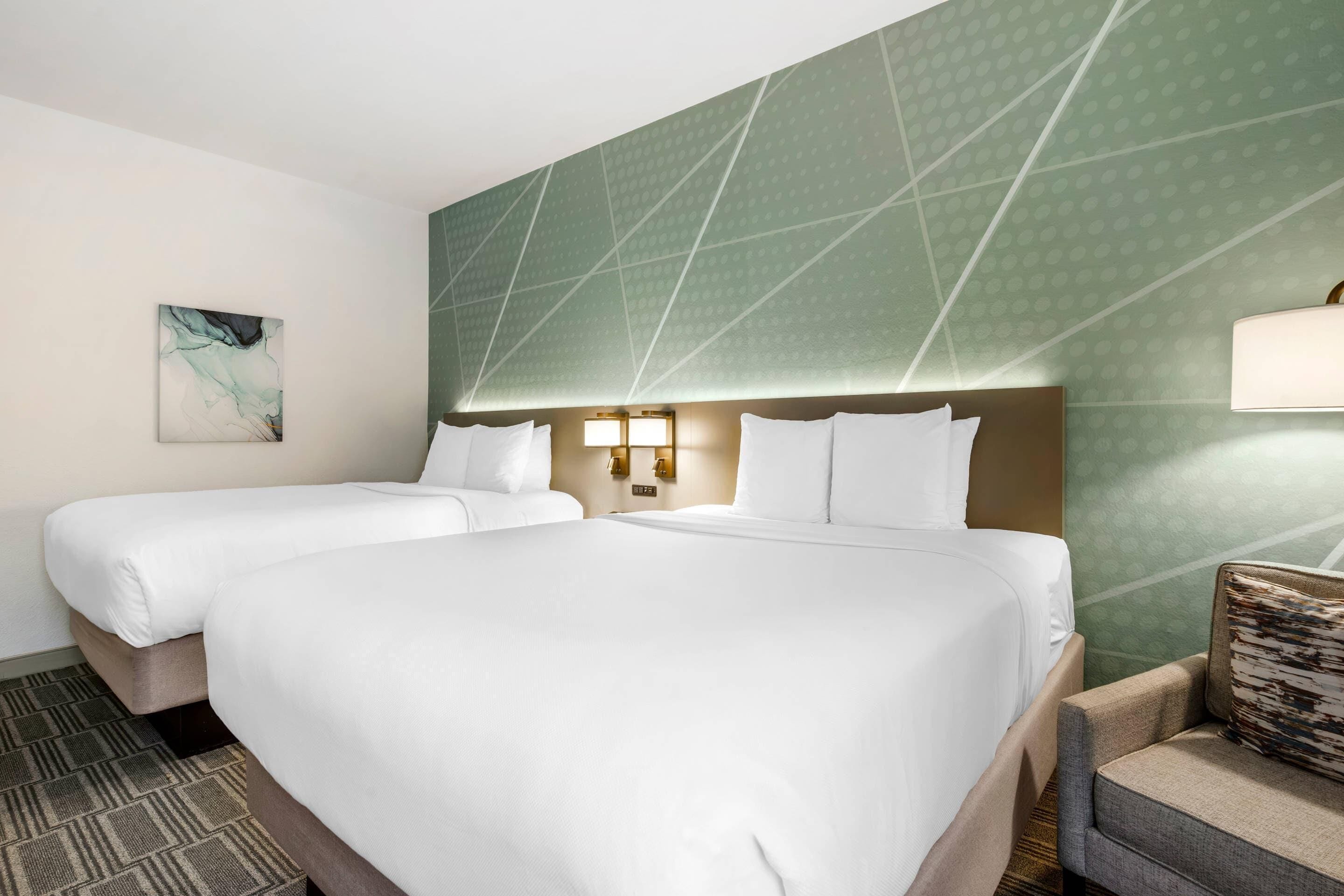 Una habitación de hotel con una cama grande, dos camas individuales, un papel tapiz estampado, un cuadro y una mesita de noche con una lámpara.