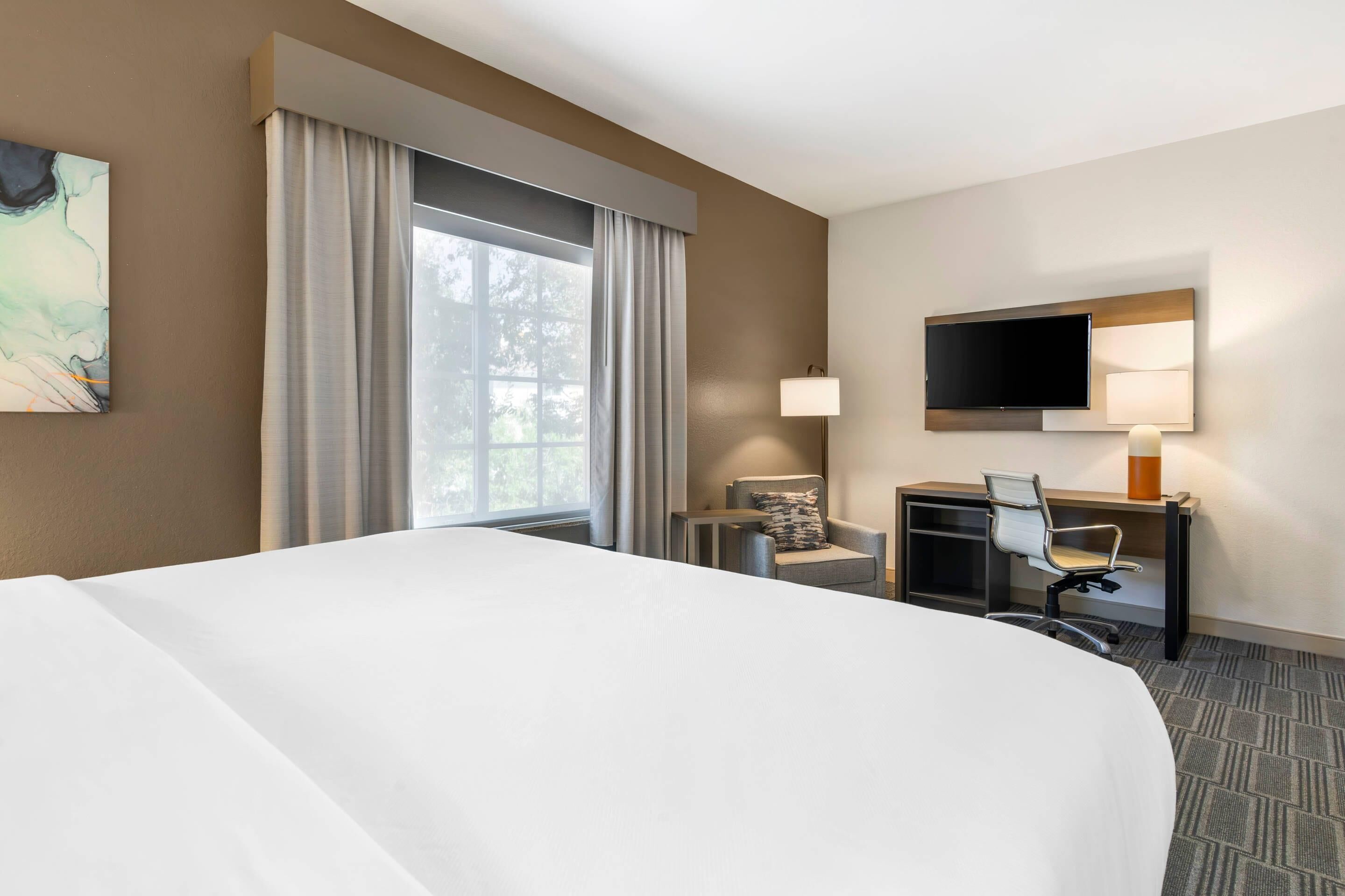 Una habitación de hotel con una cama grande, un escritorio con silla, un televisor y una ventana con cortinas.