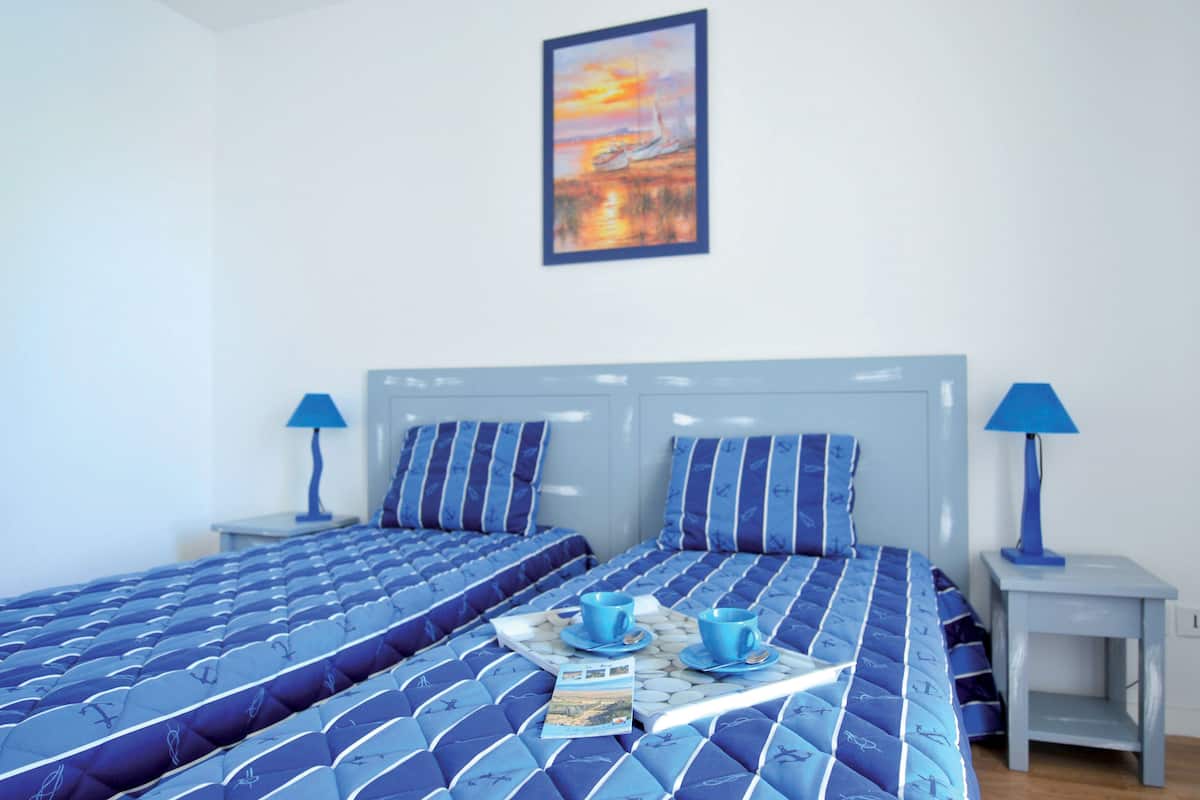 Ein Hotelzimmer mit zwei Betten, beide mit blauer Bettwäsche und passenden Kissen. Zwei Nachttische mit Lampen stehen bereit, und ein Sonnenuntergang ist als Gemälde an der Wand zu sehen.