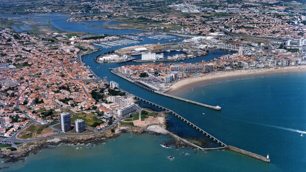 An aerial view of a coastal city with a prominent marina, residential areas, and a bridge connecting the mainland to an island. - Résidence Les Jardins de l'Amirauté - Vacancéole (Les Sables-d'Olonne)