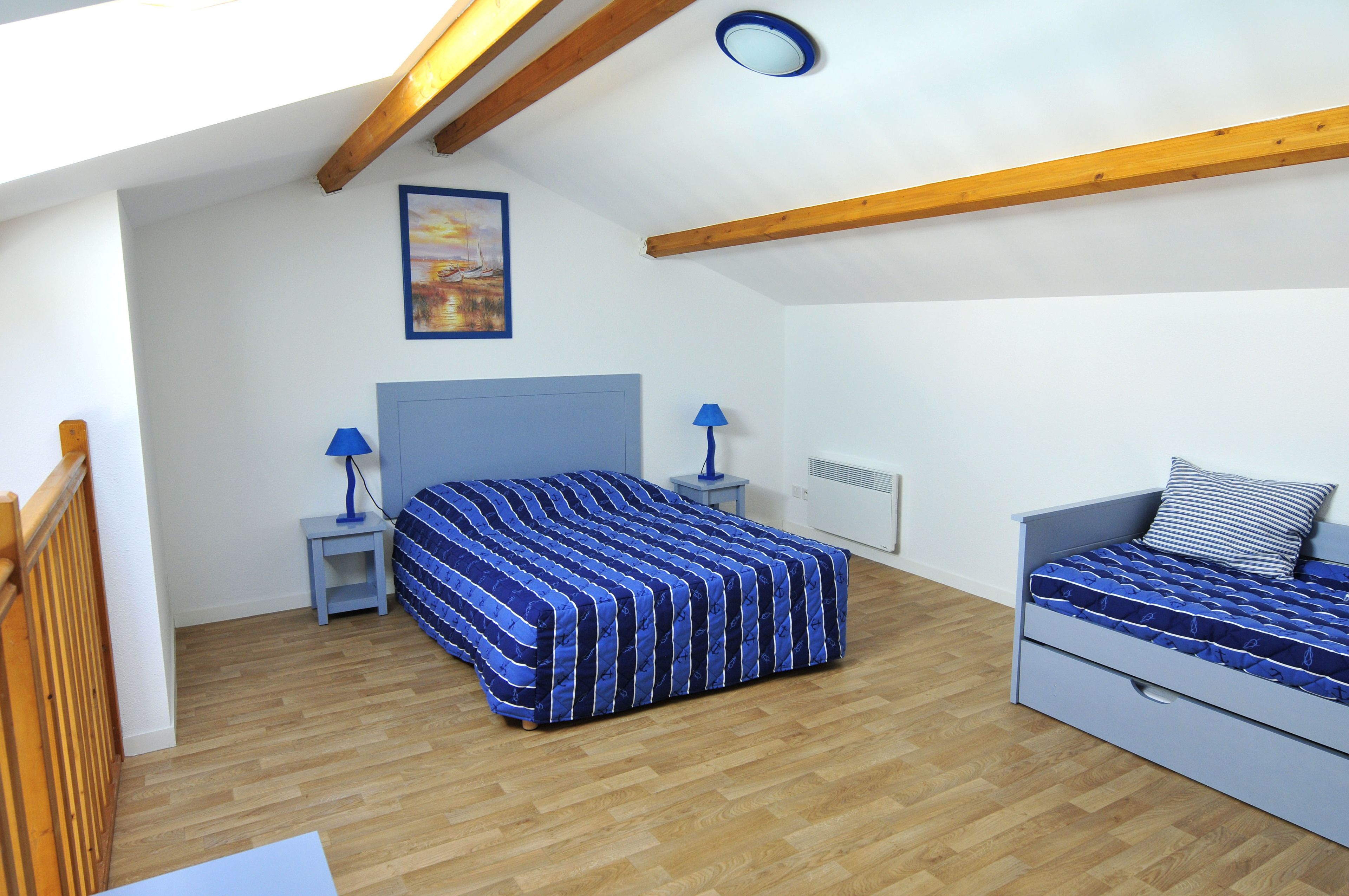 Une chambre avec un grand lit, un plus petit lit, deux tables de chevet avec des lampes, un tableau au mur et des poutres en bois au plafond.