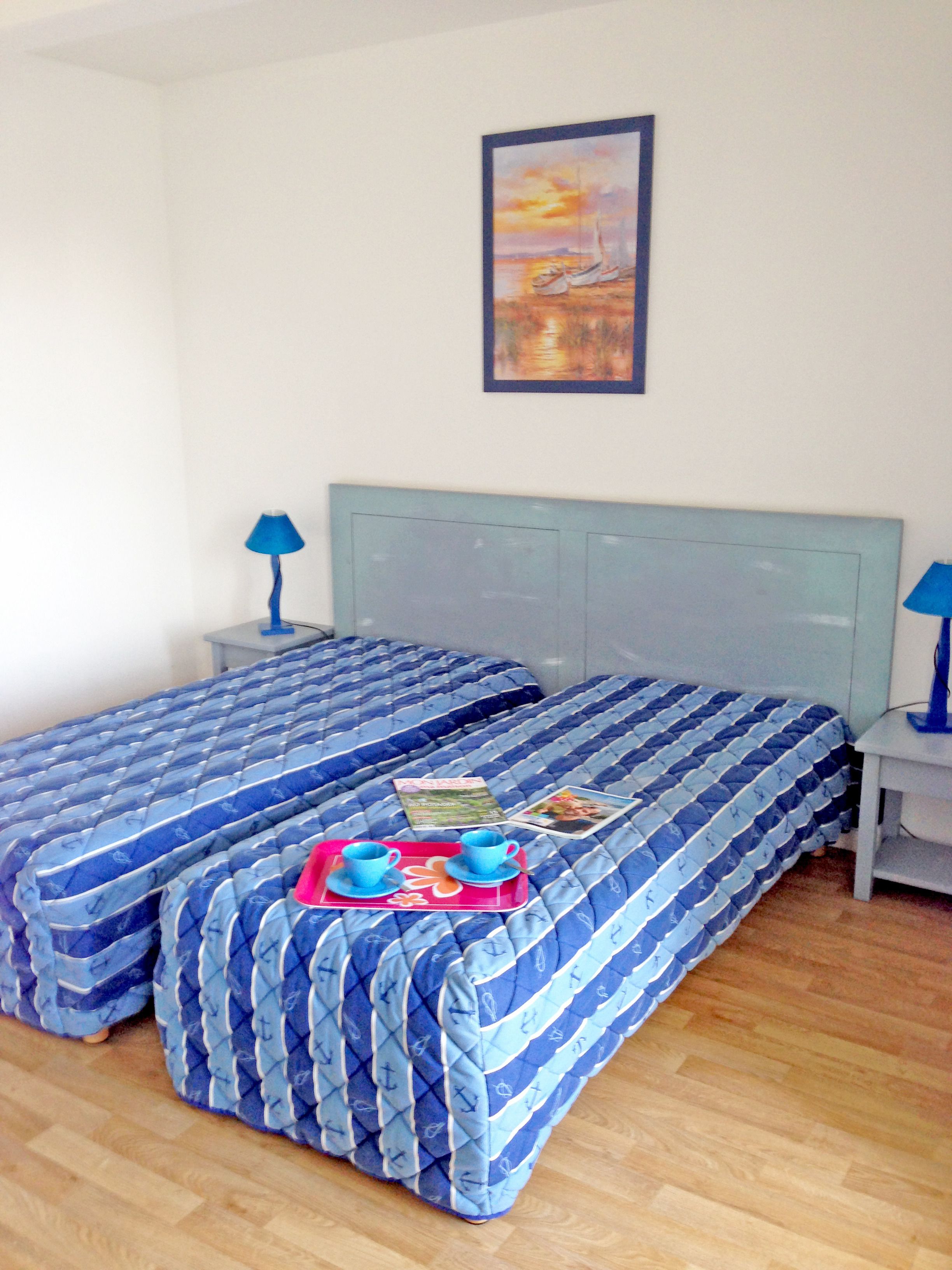A double bed with blue bedding, a small table with a tray of cups and a magazine, and a painting on the wall.