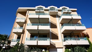 Un edificio de varios pisos con balcones, un cielo azul despejado y algo de vegetación en la base.