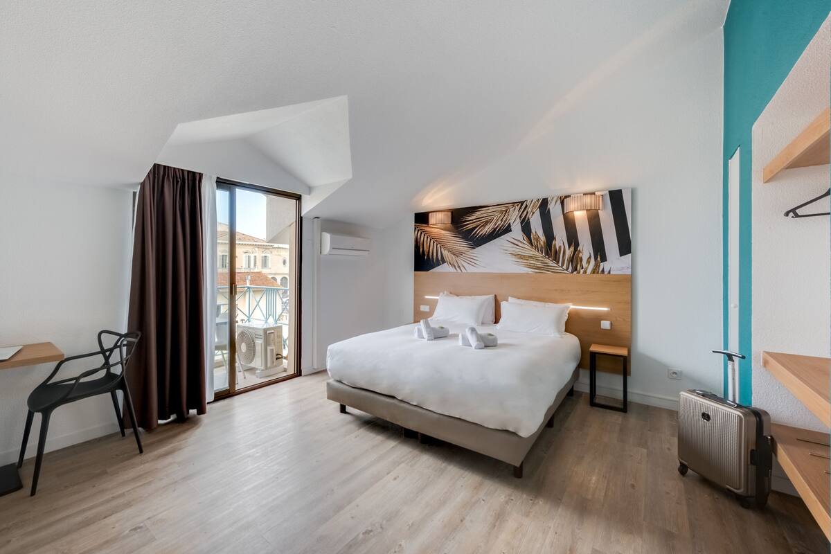 Une chambre d’hôtel moderne avec un grand lit, un bureau, une chaise, une valise et une vue sur les bâtiments, visible à travers une porte vitrée.