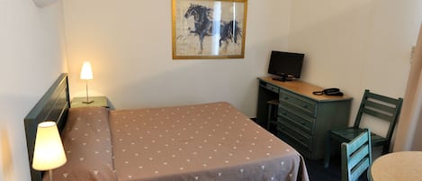 Habitación de hotel con cama, lámpara de noche, escritorio con computadora, silla y un cuadro en la pared.