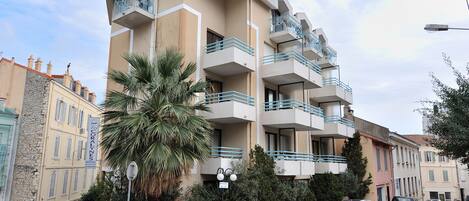 Un hotel de varios pisos con balcones, coches aparcados y una palmera al frente.