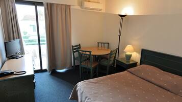 A hotel room with a bed, a desk with a TV, a chair, a table, and a lamp.
