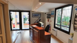 Zona de recepción con un mostrador de madera, una computadora y una silla. Hay puertas de vidrio que dan al exterior y una ventana con vista a la vegetación.
