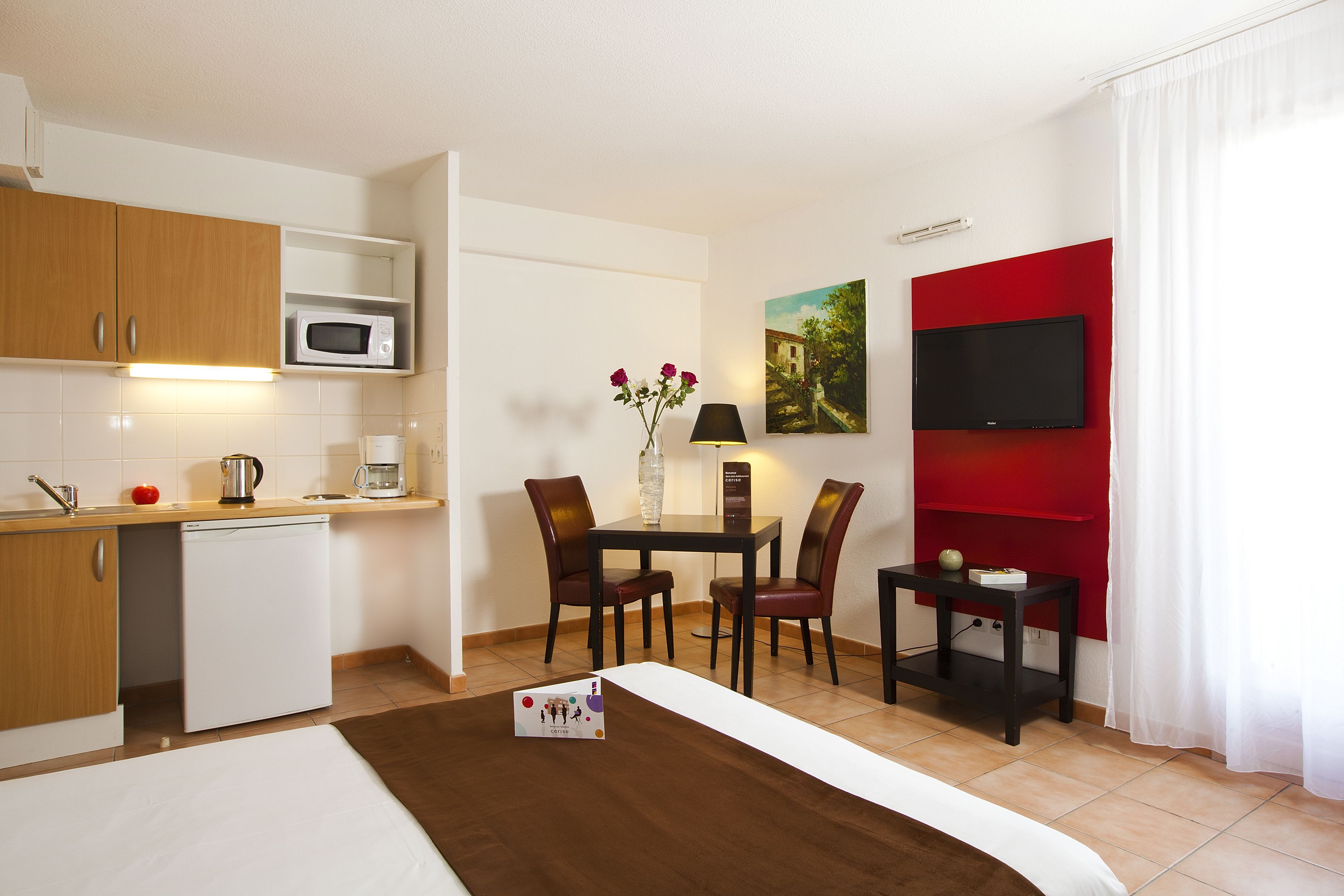 Habitación de hotel con kitchenette, comedor con mesa y sillas, cama, televisor montado en una pared roja y ventana con cortinas blancas.
