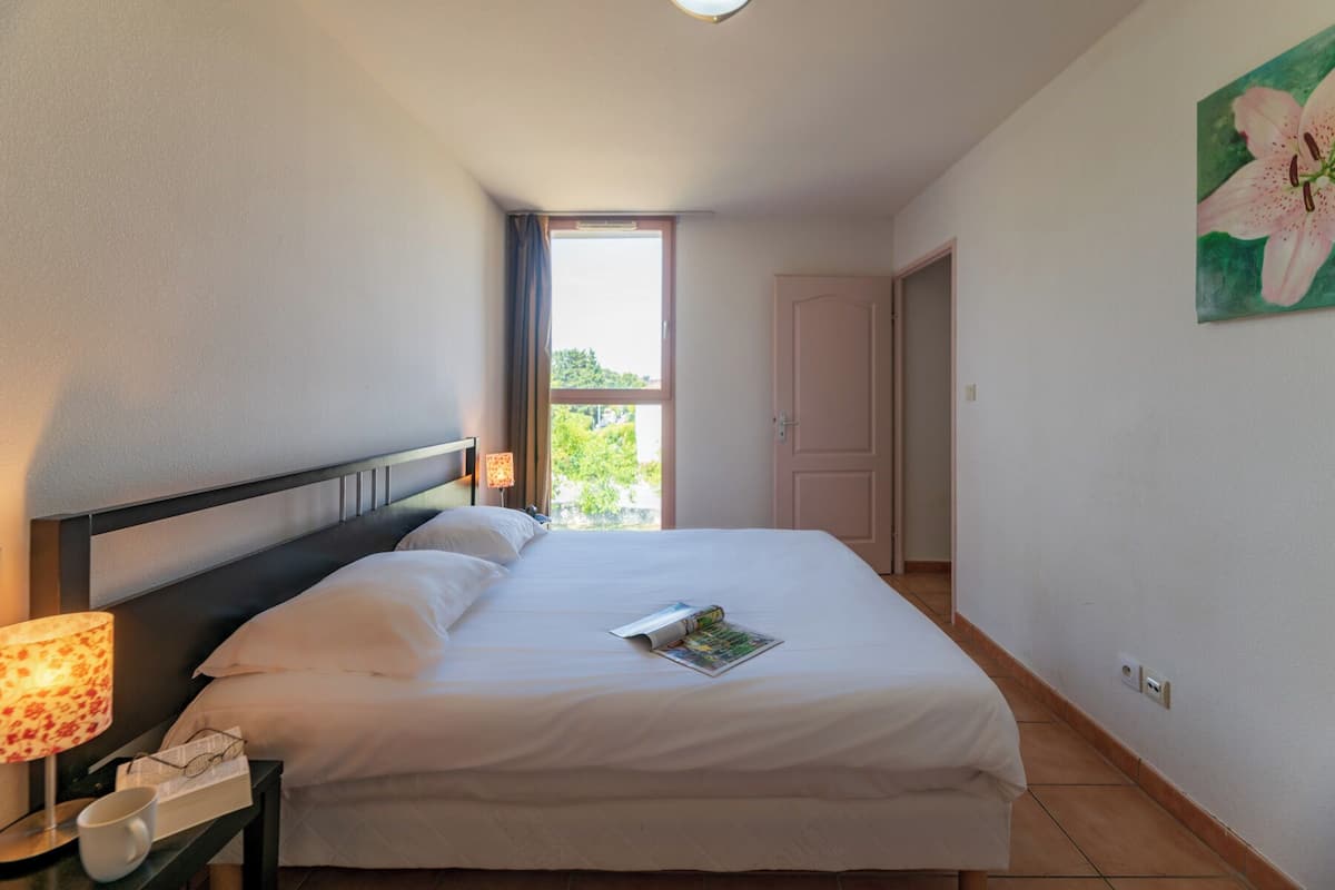 Ein ordentlich bezogenes Bett mit weißen Leinen, eine Lampe auf einem Nachttisch und ein Fenster mit Vorhängen.