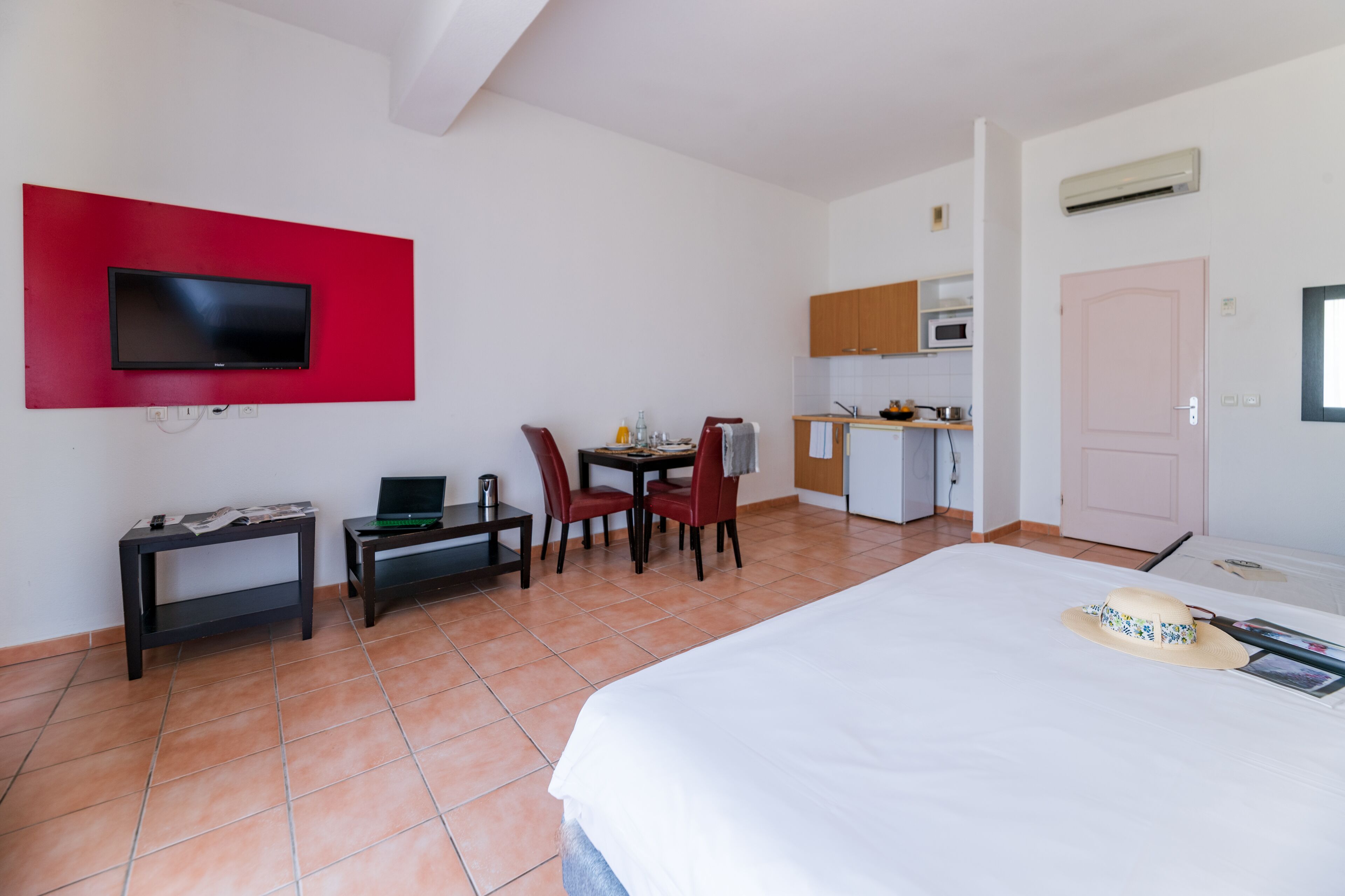 Habitación de hotel con cama, escritorio con una computadora portátil, televisión montada en una pared roja, kitchenette y una puerta.