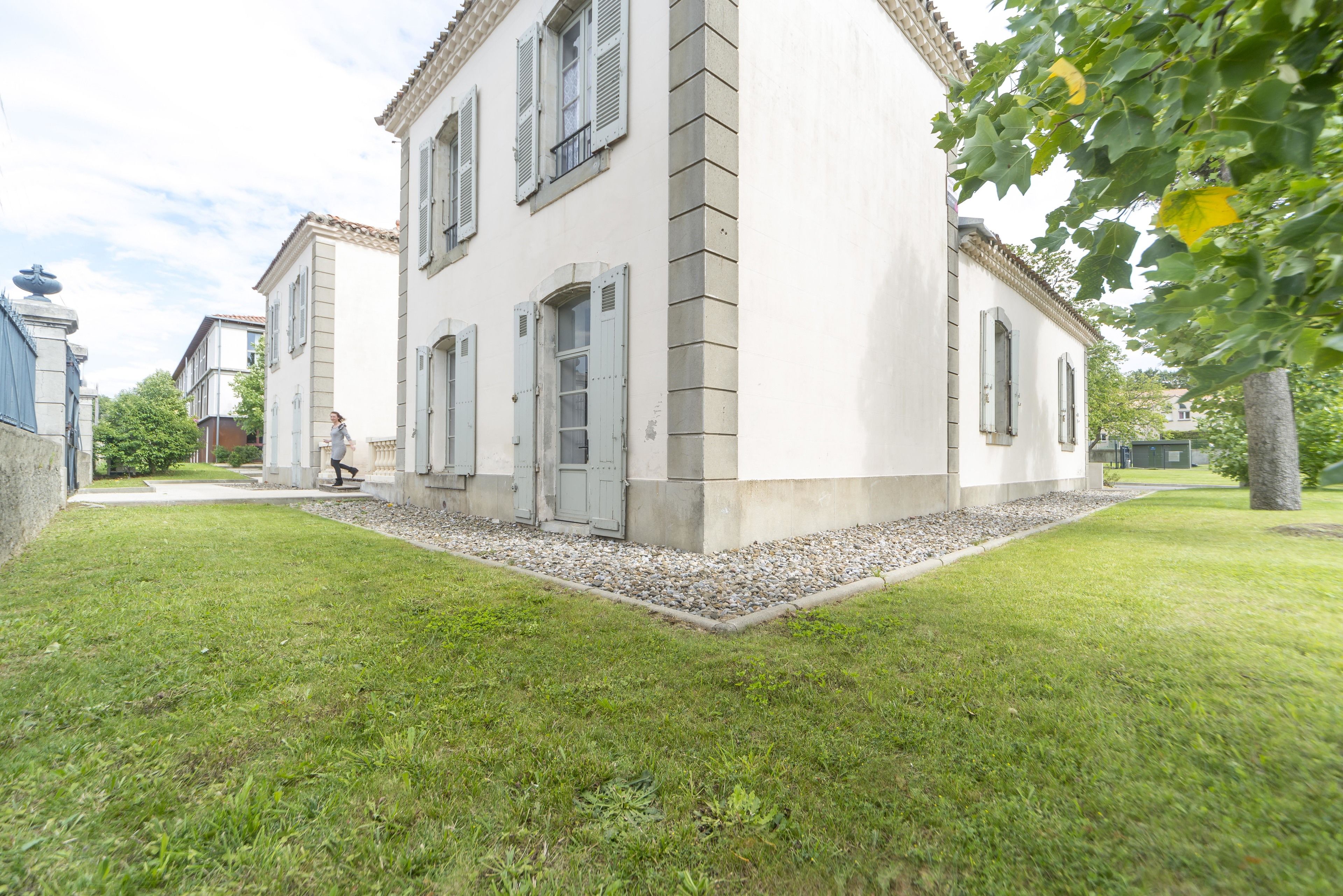 A two-story building with a white facade and grey shutters, surrounded by a well-maintained lawn and a tree with yellow flowers.